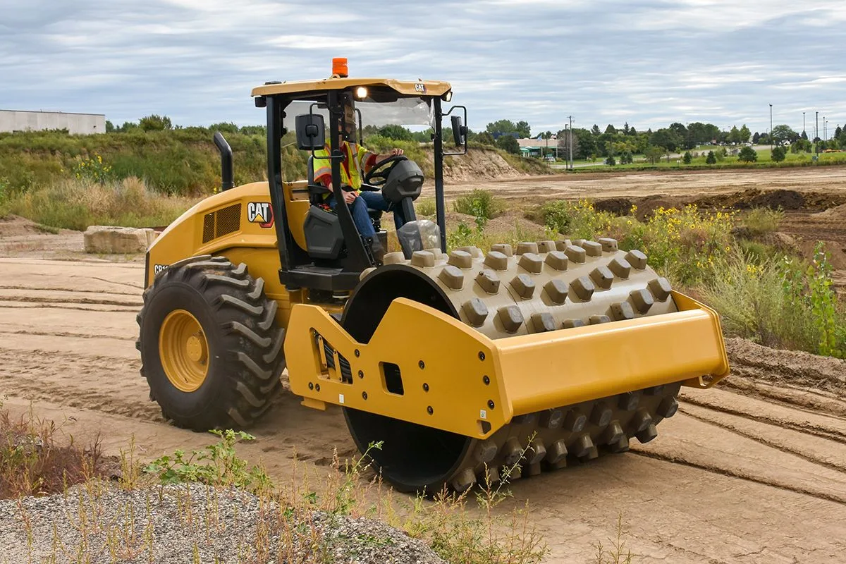 Padfoot roller compacting loose dirt across a wide, freshly graded building site with a low pile of dirt and trees in the distance