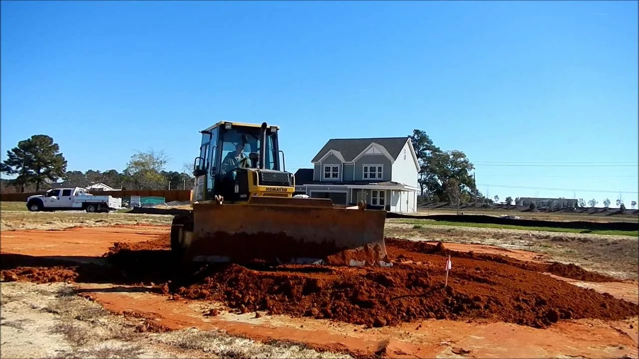 Dozer pushing a pile of red clay across a new house pad, with a newly built two story house and a pickup truck behind it