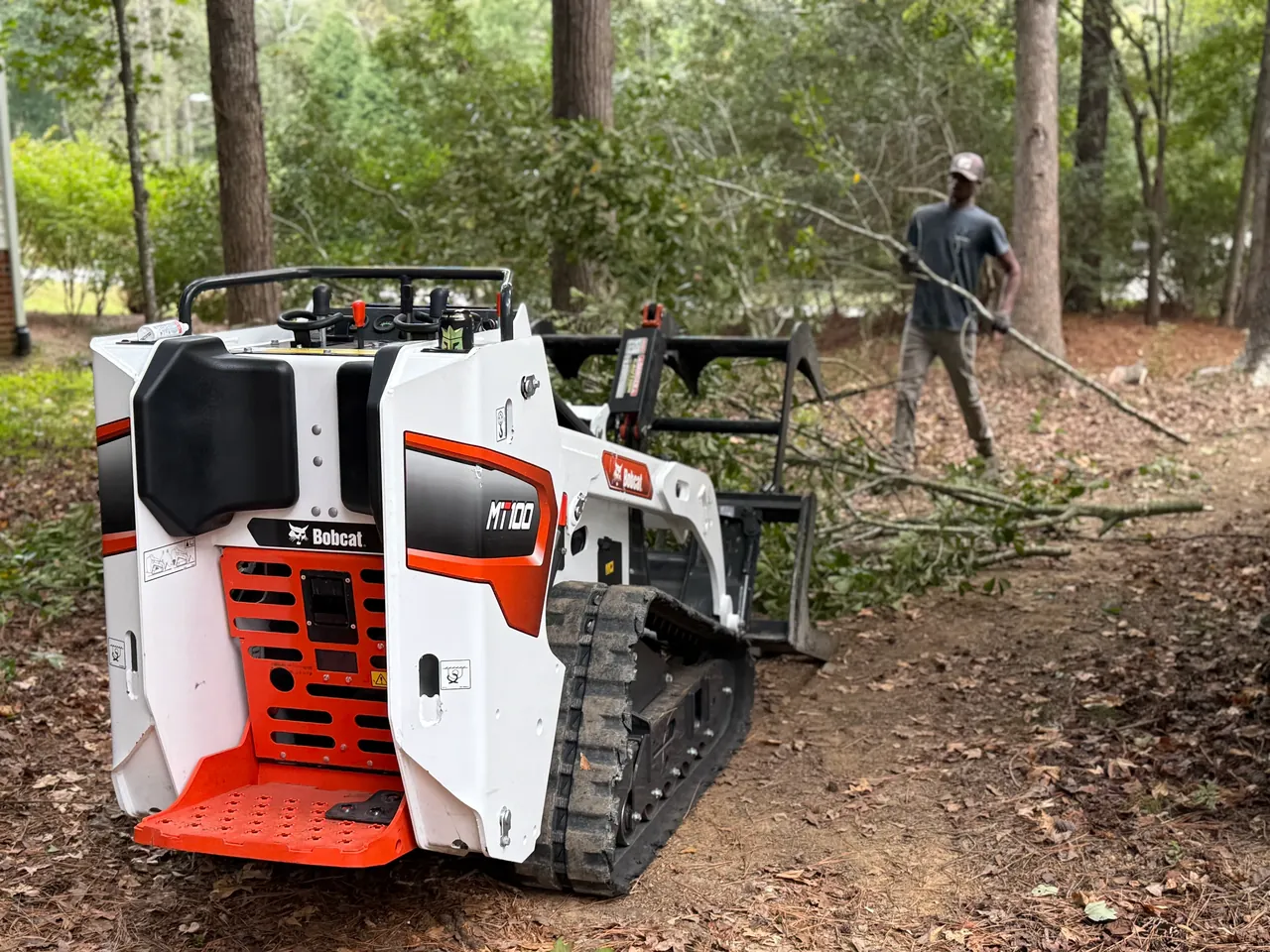 Compact track loader with a brush grapple beside cut limbs while a worker drags a branch across a wooded lot